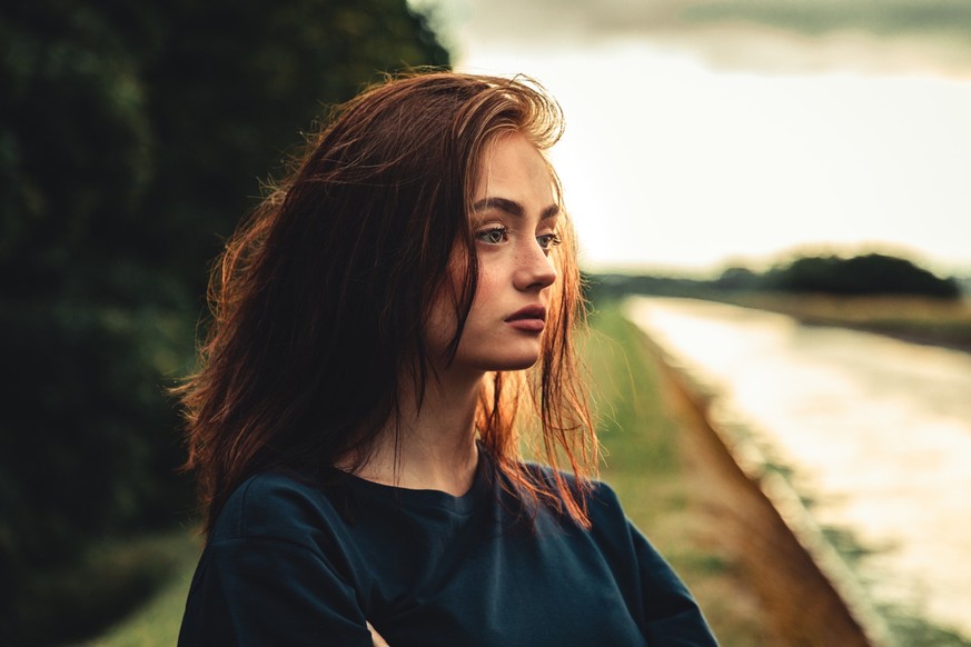 Young day dreaming, pensive and sad looking young woman standing alone outdoors close to river in sunset twilight. Thinking, looking to the horizon. Real People Youth Culture Lifestyle Portrait.