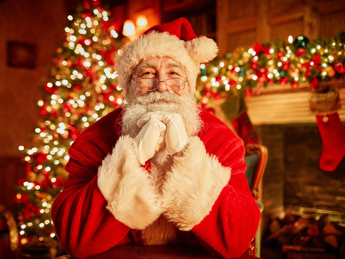 Front view portrait of traditional Santa Claus smiling at camera resting head on arms with christmas tree in background