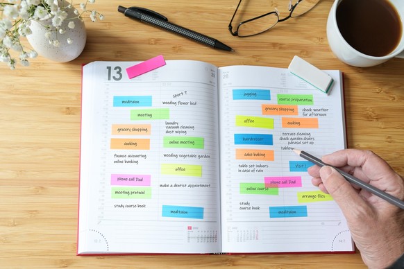 Hand of a woman writing appointments in a diary organizer or schedule calendar with a lot of colorful sticky notes for her time management on a wooden desk with coffee, flowers and glasses, selected f ...