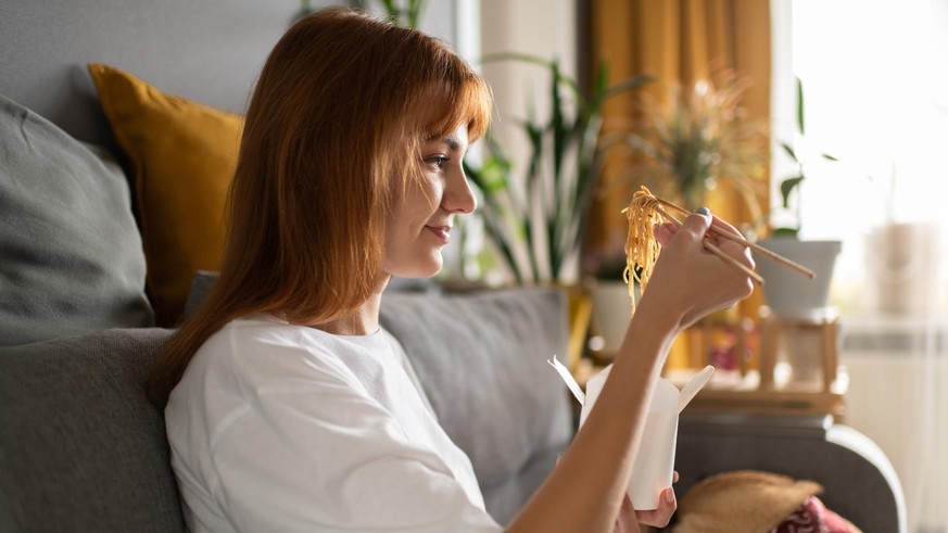 Side view of glad female eating Chinese food while sitting on couch near dog in cozy living room