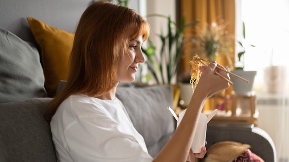 Side view of glad female eating Chinese food while sitting on couch near dog in cozy living room