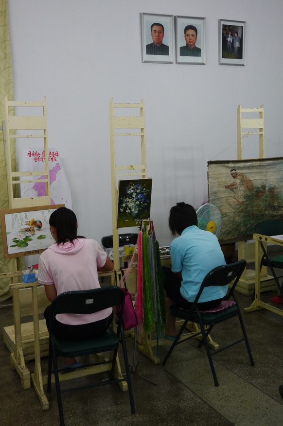 Pyongyang, North Korea - August 18, 2012: Two young women working in an embroidery workshop in Pyongyang, North Korea.On the wall there are pictures of the former politcal leaders Kim Il-sung and Kim  ...