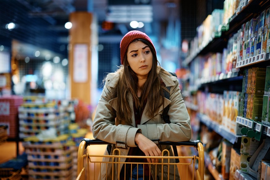 Young sad woman with empty shopping cart among produce aisle at supermarket.