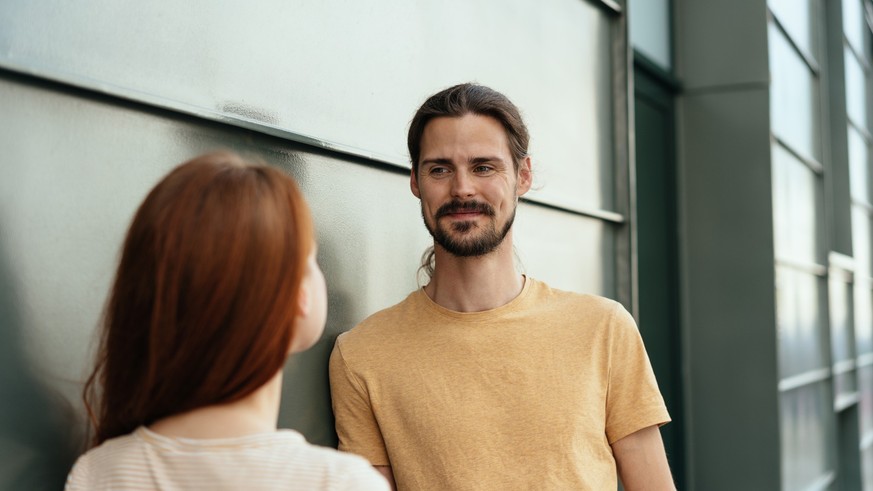 Attractive man chatting to a young woman in an over the shoulder view as he smiles quietly at her as they stand against a grey exterior wall
