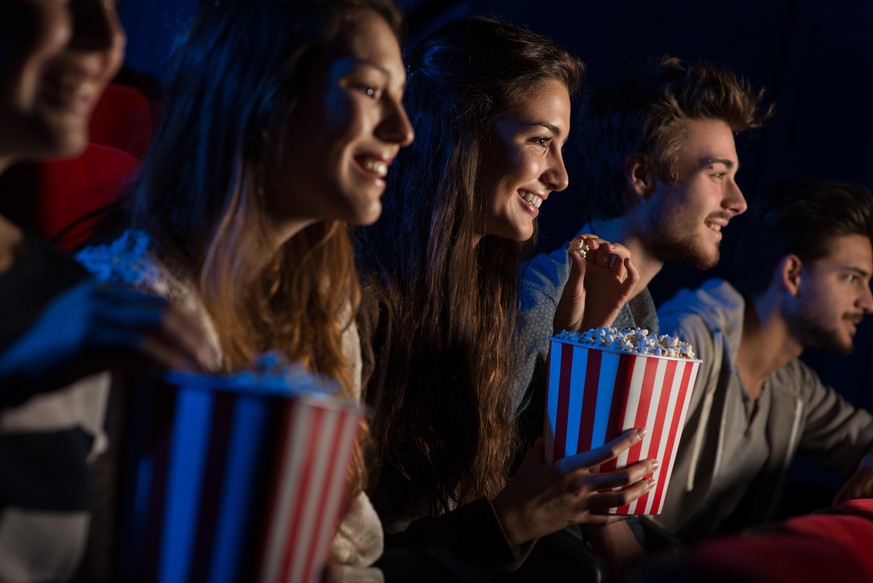 Group of teenager friends at the cinema watching a movie together and eating popcorn, entertainment and enjoyment concept