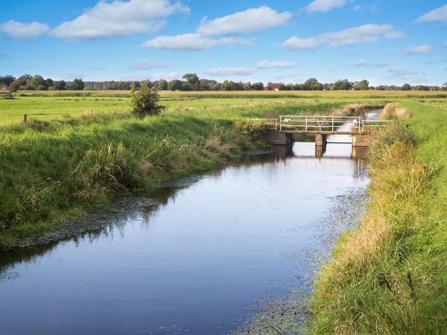 Landscape view along a drainage ditch (Schloot) with a small pedestrian bridge between green meadows in front of a building Sky with small clouds in northern Germany.