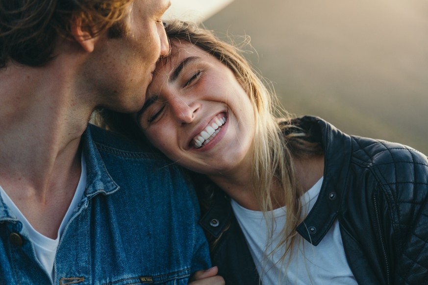 Close-up of romantic young boyfriend kissing smiling beautiful girlfriend on forehead while enjoying weekend at sunset