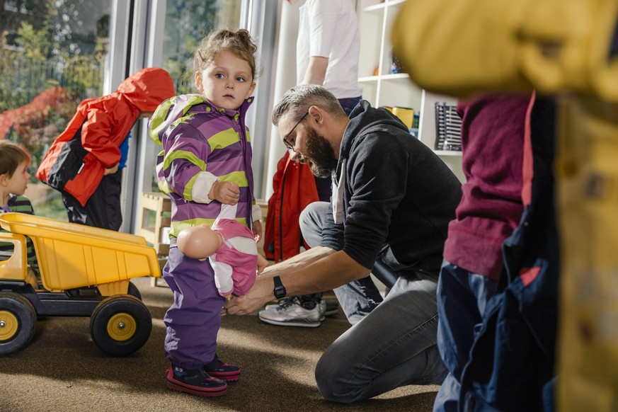 Pre-school teacher helping girl putting on rainwear in kindergarten model released Symbolfoto property released PUBLICATIONxINxGERxSUIxAUTxHUNxONLY MFF04109