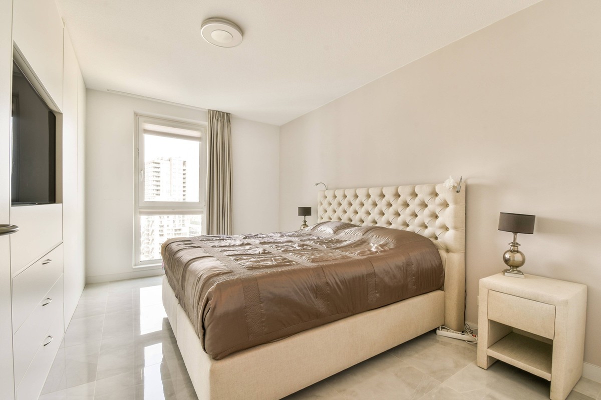 Modern minimalist bedroom showcasing a plush bed, elegant bedside lamps, and soft beige tones. Natural light spills through a large window, creating a serene atmosphere Model Released ProCreators_ID28 ...
