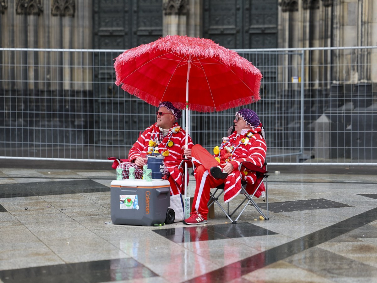 27.02.2025, Nordrhein-Westfalen, Köln: Zwei Karnevalisten sitzen bei regnerischem Wetter unter einem Sonnenschirm auf der Domplatte. Am Donnerstag beginnt mit der Weiberfastnacht die Karnevalssession ...