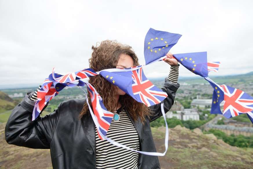 Brexit - Young adult woman holding European Union and British Union Jack flags on a windy hilltop. Arthur&#039;s Seat, Edinburgh, Scotland.