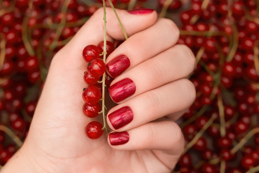 Female hand with white red design. Glitter red nail polish manicure. Woman hand hold red current berries on red current background