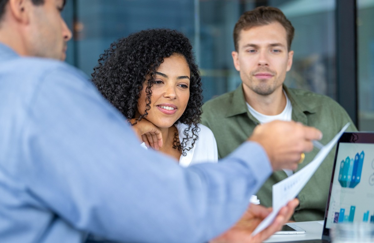 Couple listening to a businessman, banker or financial advisor who has a laptop computer showing financial charts and graphs. He is showing them paperwork contract or information.