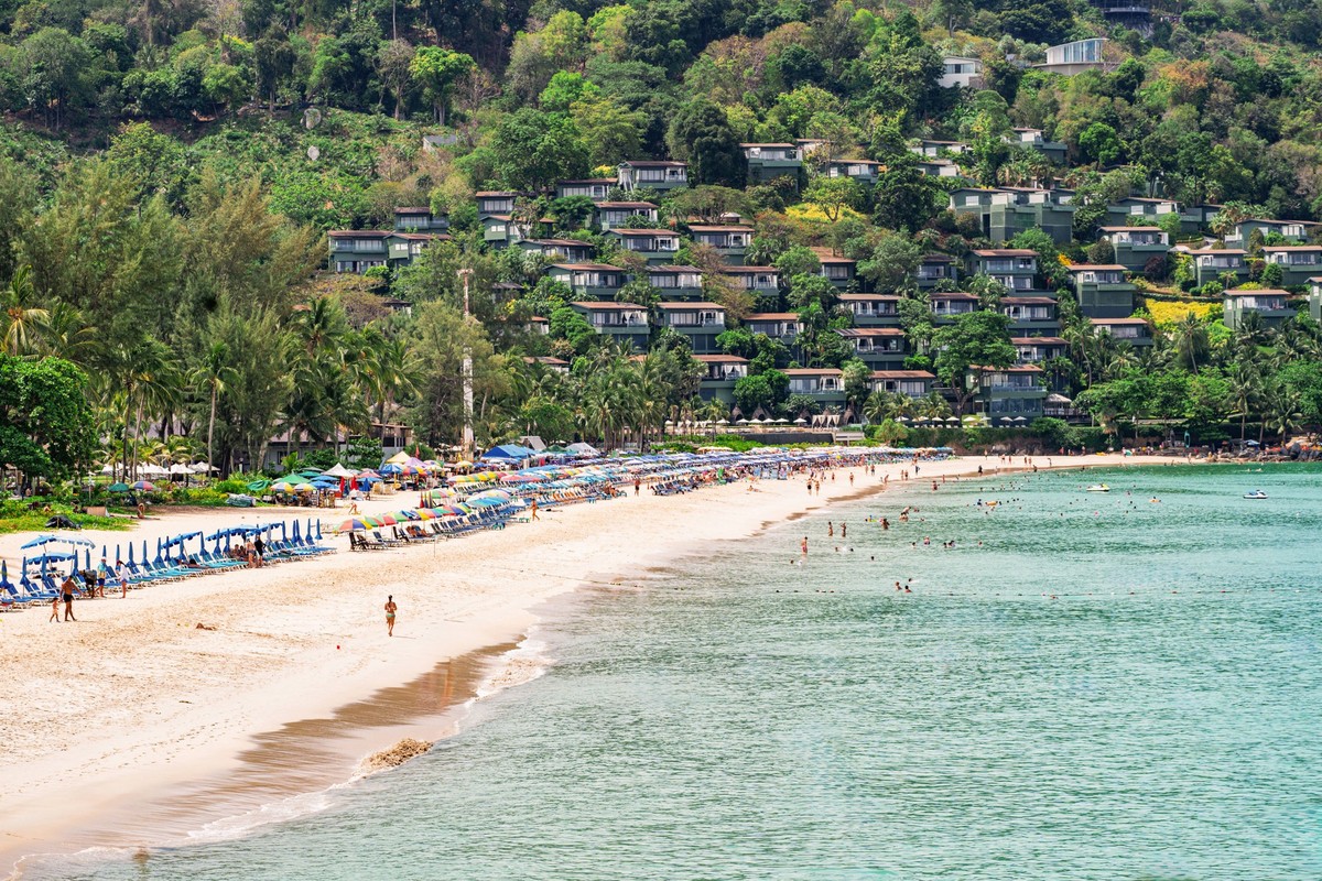 A scenic beach in Phuket, Thailand, lined with umbrellas and sunbeds, bustling with people enjoying the sun, and hillside villas overlooking the turquoise sea and lush greenery Model Released IrinaChe ...