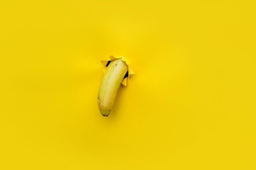 A small banana curved downward through a torn hole in yellow paper. Tropical fruit, vegetarianism. Bright background with copy space.The concept of impotence, erectile dysfunction, joke.Half past five