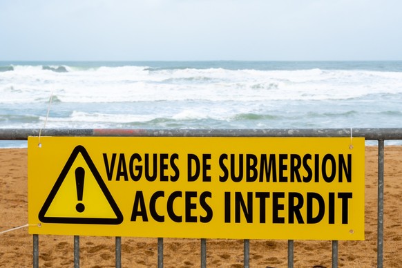 Weather alert - Storm warning: Yellow warning sign on a metal barrier prohibiting access to the beaches of the Atlantic coast with the message &quot;Flood waves - Access prohibited&quot; written in Fr ...
