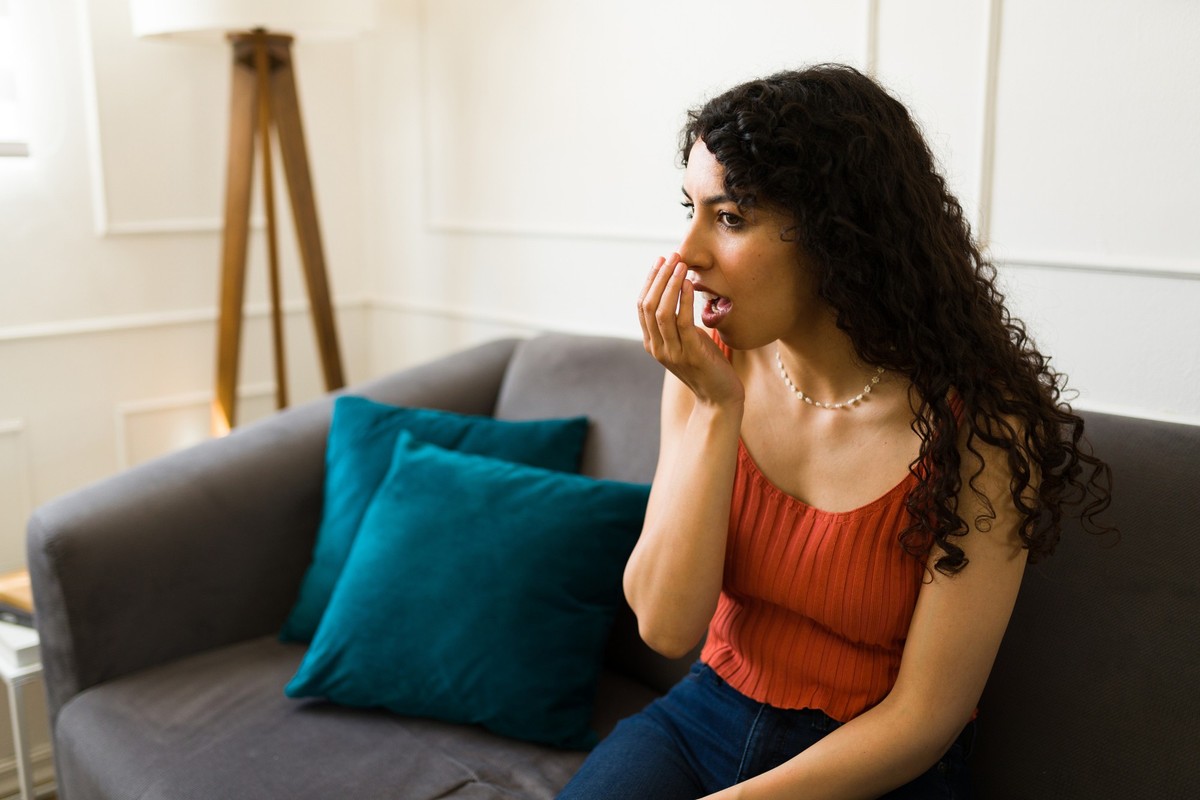 Attractive young woman with health problems checking her bad breath and suffering from halitosis while sitting on the sofa at home