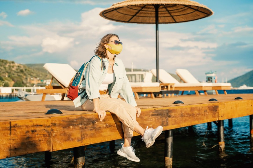Young woman with a mask on her face sits on a pier by the sea, summer travel trend 2020, health safe vacation