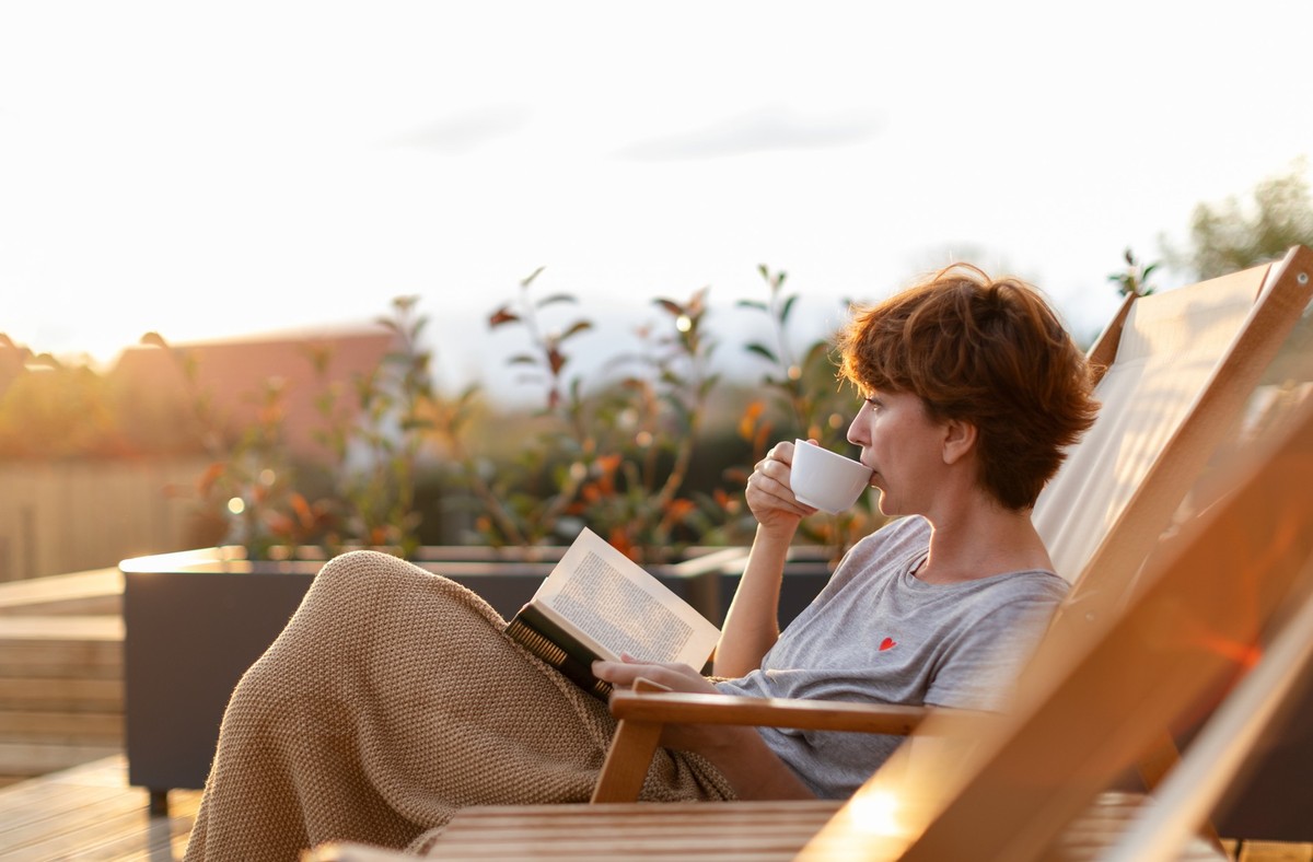 Beautiful woman is sitting on patio on the deck chair and enjoying beautiful surrounding while drinking coffee or tea and reading a book