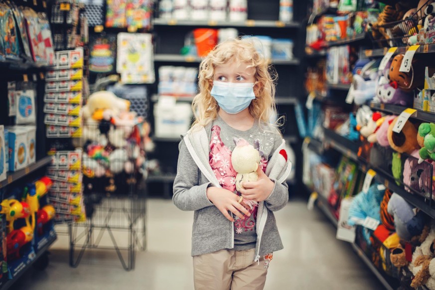 A new normal. Caucasian blonde girl in sanitary face mask shopping at toy store. Child wearing protective mask against coronavirus. Safety, health protection during covid-19 quarantine.