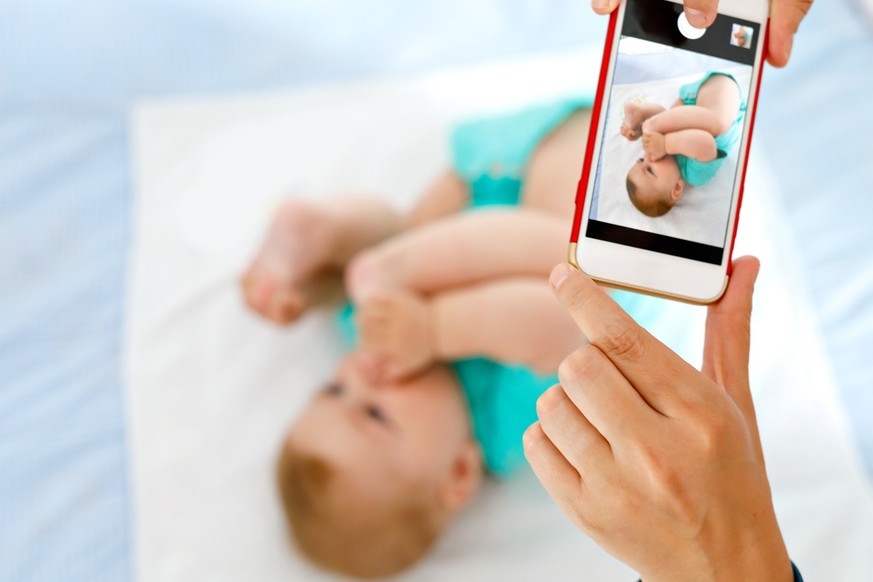 Parent taking photo of a baby with smartphone. Adorable newborn child taking foot in mouth. sucking feet. Digital family memories