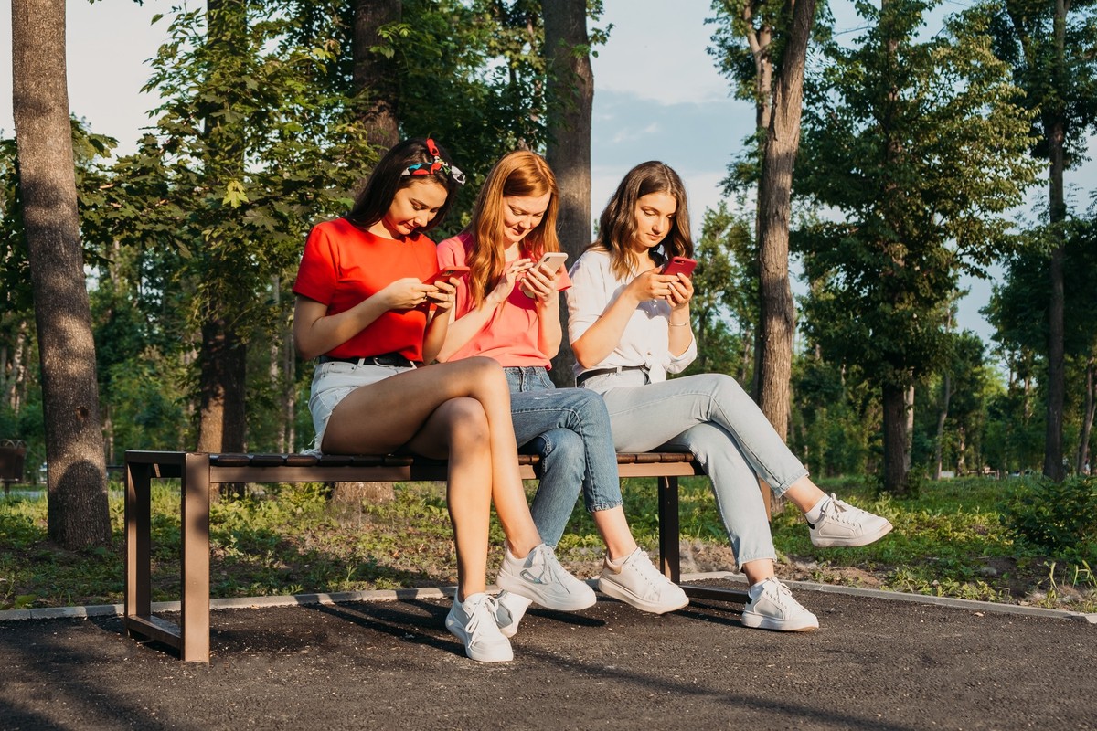 Three girls chatting with their smartphones at the park. Gen Z young girl friends using gadget and having fun outdoors model released