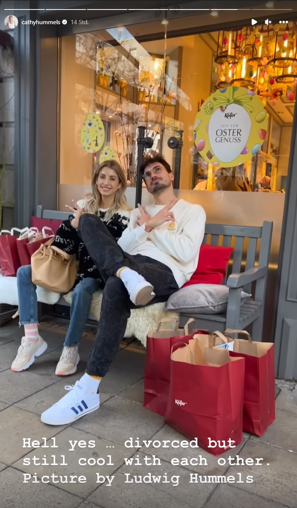 Cathy und Mats Hummels sitzen zusammen auf einer Bank und machen mit den Armen Kreuz-Geste.