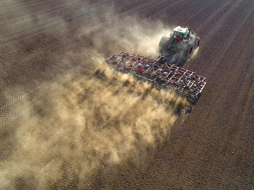 Big farm tractor tilling dusty Springtime fields, aerial view.