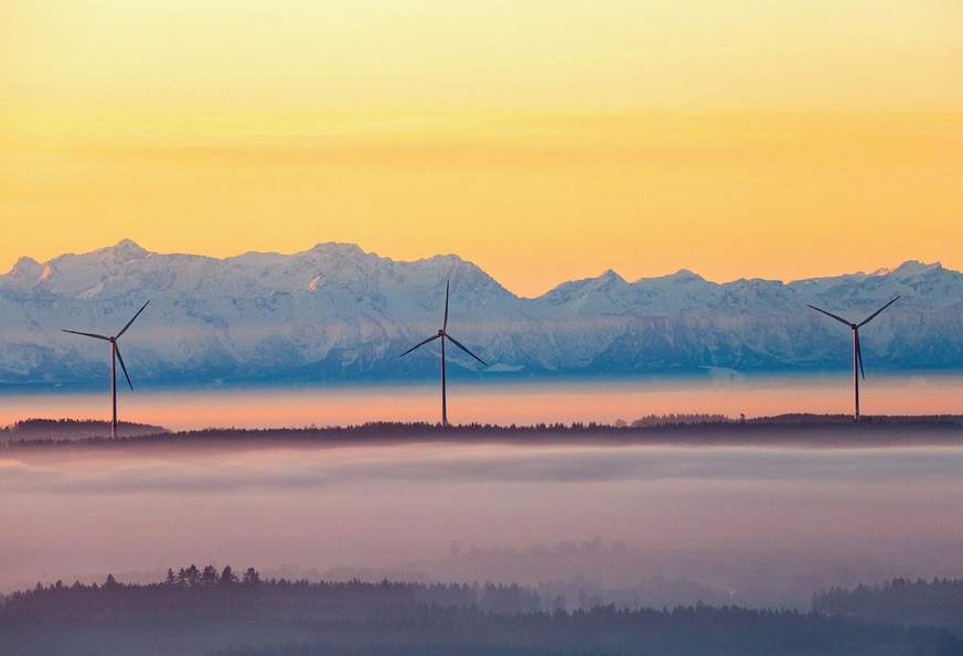 17.12.2023, Baden-Württemberg, Uttenweiler: Drei Windräder ragen am Morgen kurz nach Sonnenaufgang aus der Nebeldecke, während im Hintergrund durch den Föhn die über einhundert Kilometer entfernten Al ...