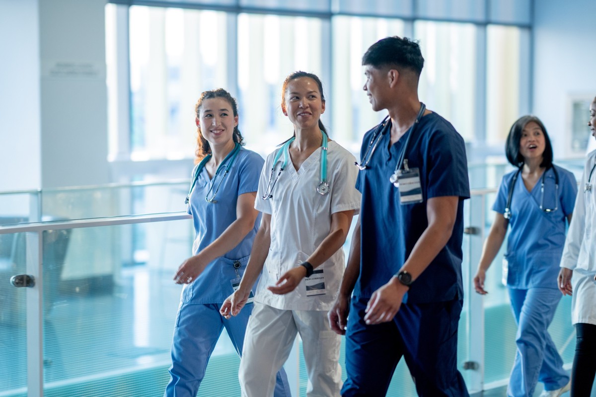 A small team of medical professional&#039;s are seen walking through the halls of a hospital as they make rounds together to patients. They are each dressed in scrubs and are smiling as they talk amon ...