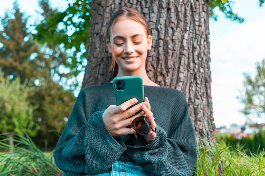 Happy smiling young woman leaning on tree in the garden, reading messages on her mobile phone, smiling happy. Young women social media lifestyle concept shot.