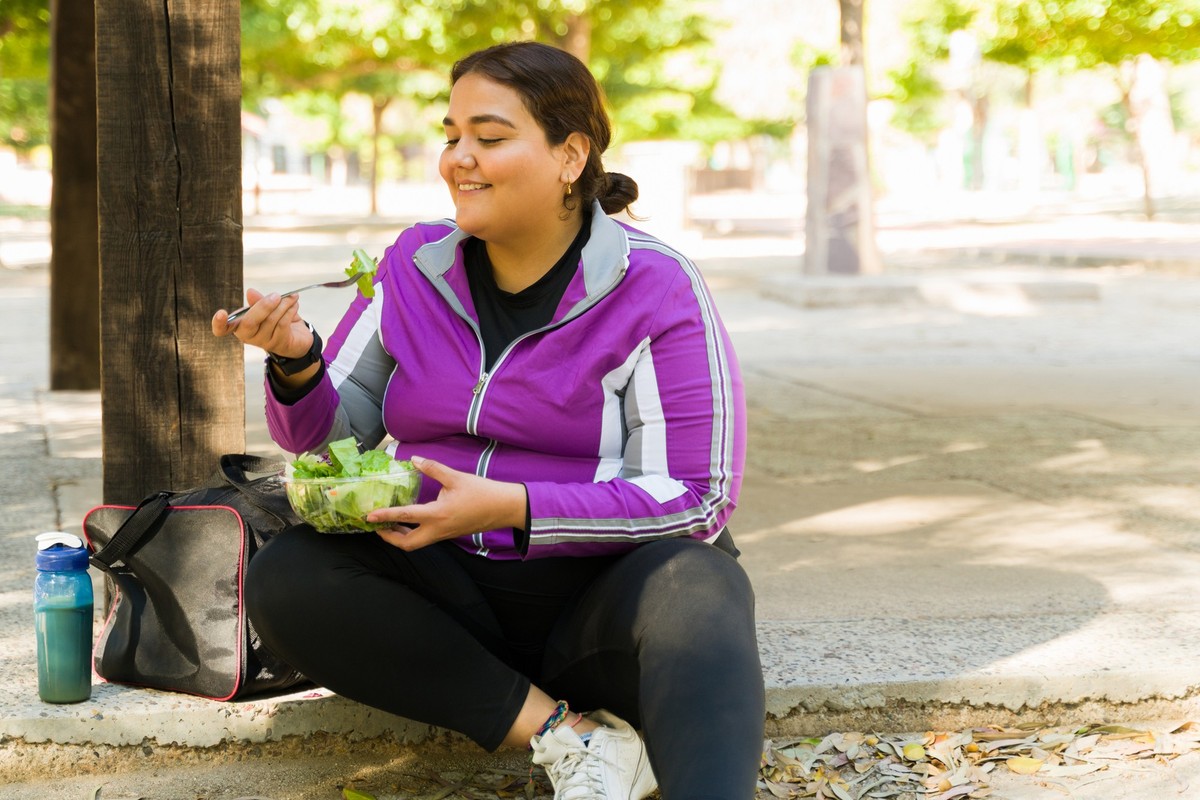 Happy overweight woman smiling while eating a healthy green salad before starting her cardio workout outdoors