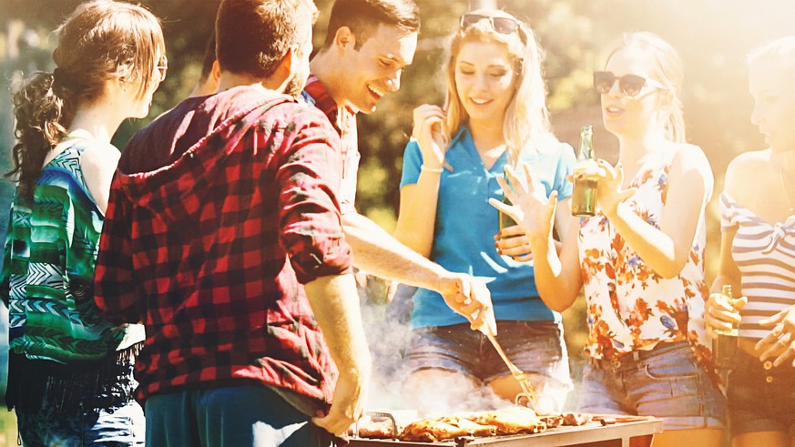 Group of mid 20&#039;s people having backyard barbecue party. There are three guys and four girls gathered around heavily smoking grill and sipping cold beer. One of the guys is being today&#039;s che ...