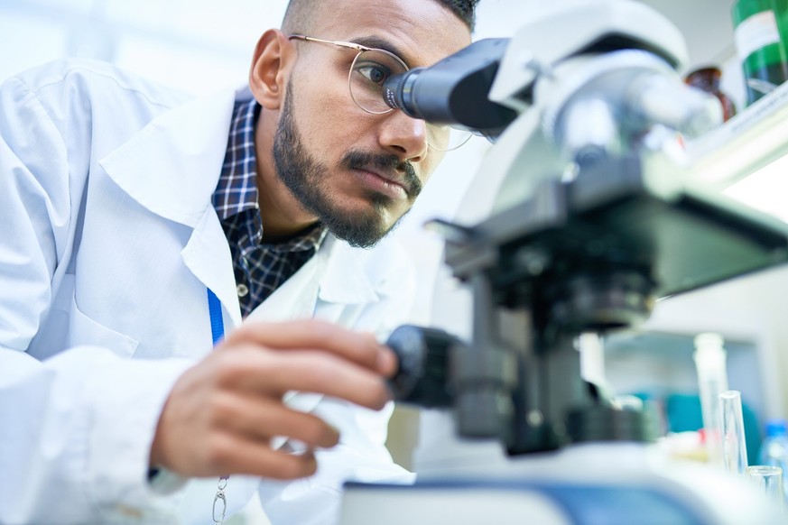 Portrait of young Middle-Eastern scientist looking in microscope while working on medical research in science laboratory, copy space