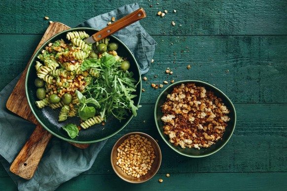 Summer vegetarian pasta salad with broccoli pesto, peas, arugula, olives, pine nuts and bread crumbs on dark green background. Top view.