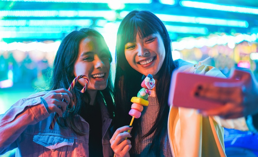 Happy asian girls eating candy sweets and taking selfie at amusement park - Young trendy friends having fun with technology trend - Tech, friendship and influencer concept - Focus on right female face