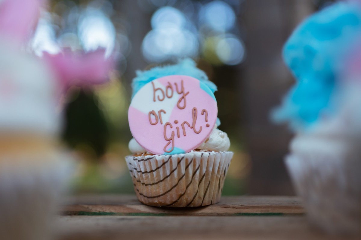 A close-up of a cupcake with a decorative topper that reads &quot;Boy or Girl?&quot; in a gender reveal party setting. The cupcake is part of a festive display, on a wooden table with other cupcakes a ...