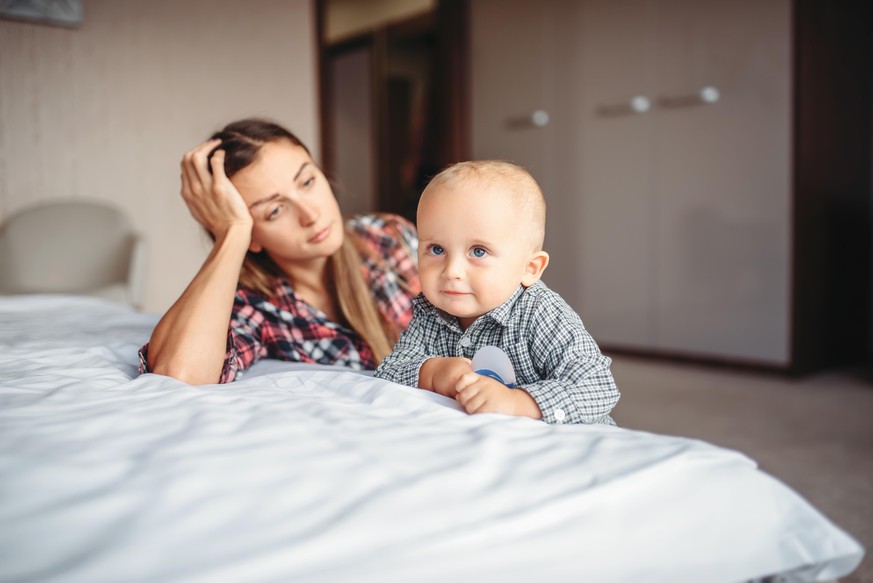 Tired mother looks on her child playing on the bed