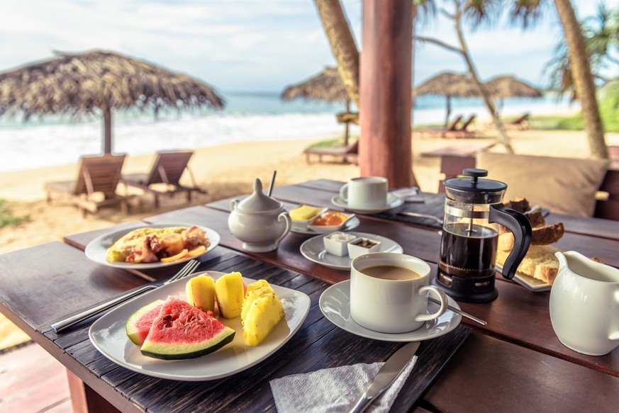 Breakfast on sea beach in Sri Lanka. Table setting with vegan food and coffee in restaurant outdoor. Buffet with exotic fruits on ocean coast. Concept of travel and vacation in tropical resort.