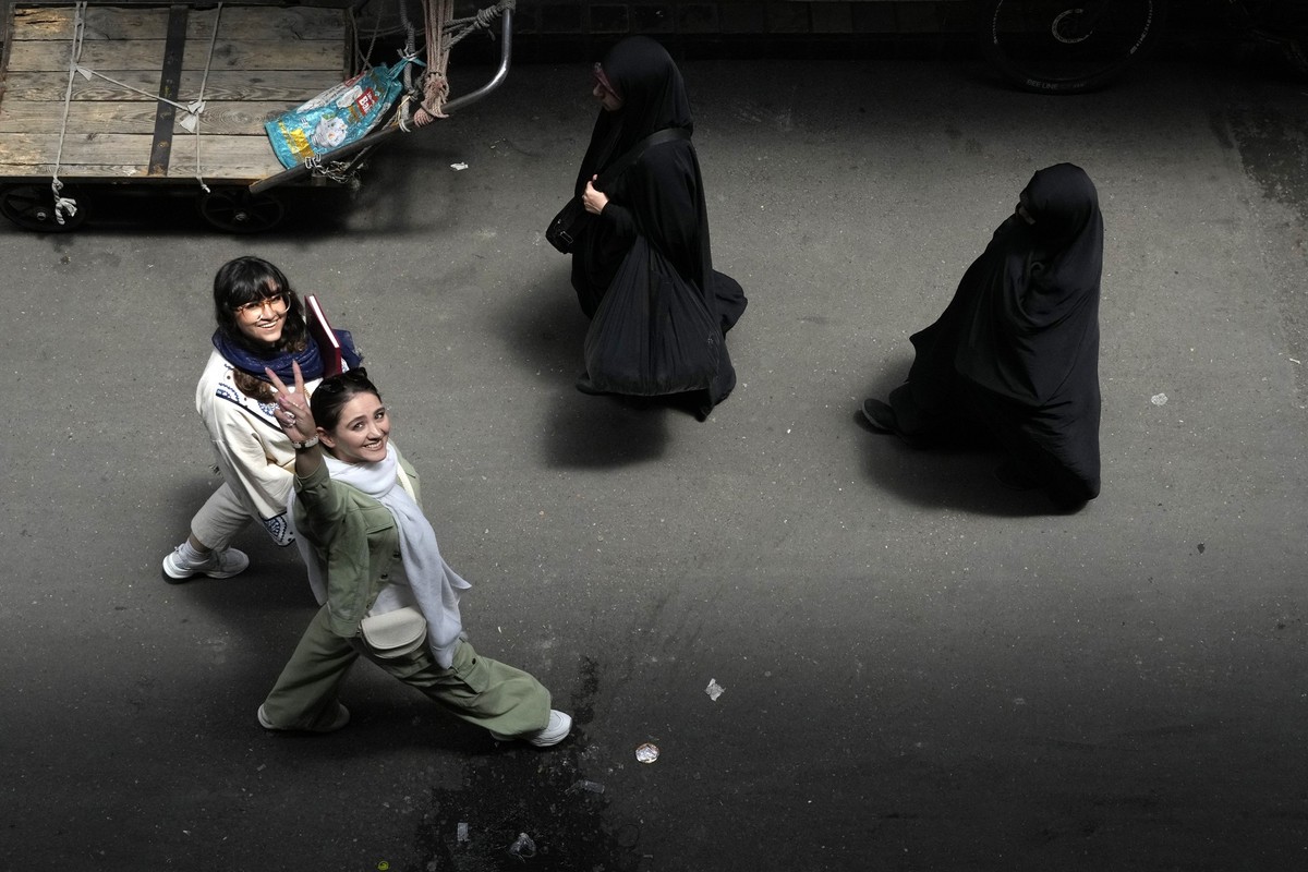 PRODUKTION - 13.06.2024, Iran, Teheran: Eine iranische Frau, die ihr obligatorisches islamisches Kopftuch nicht trägt, zeigt ein Siegeszeichen, während zwei bis zum Boden verschleierte Frauen auf dem  ...