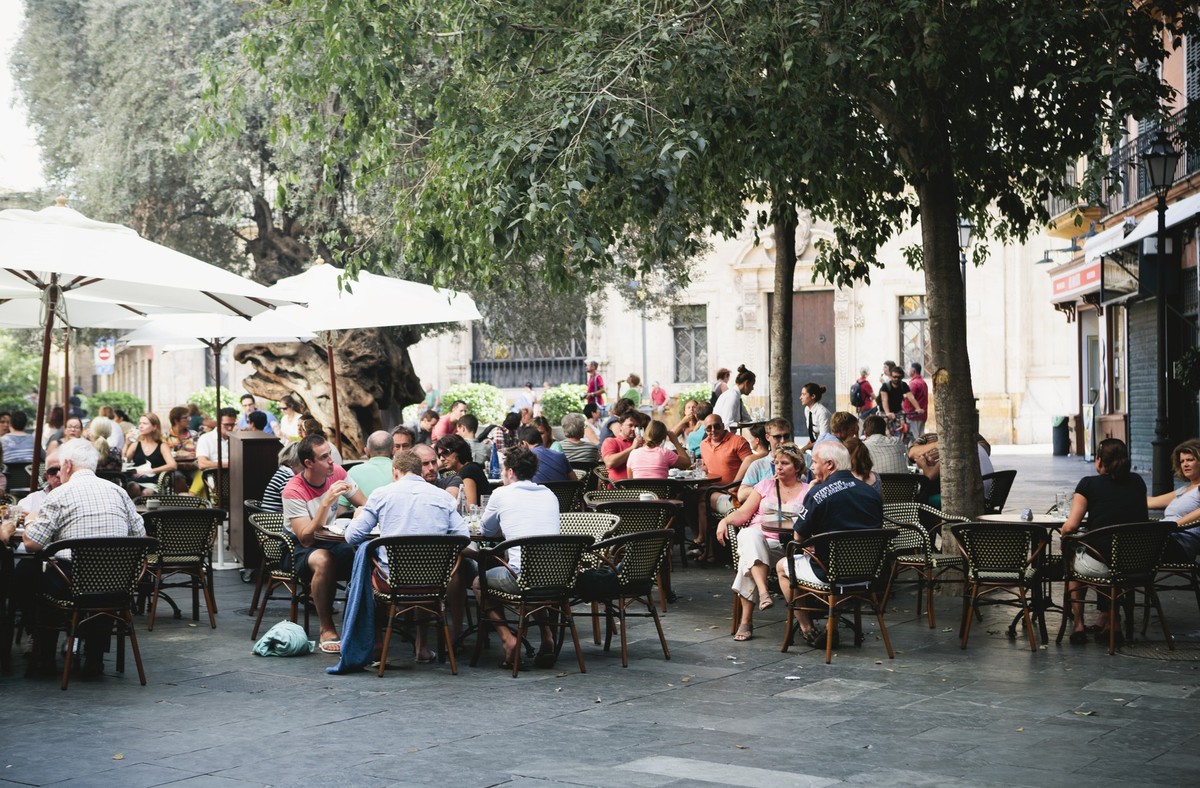 This long shot shows many diverse people sitting in an outdoor dinning area near a restaurant in Palma, Majorca. The tables are covered by tall, white, fabric, sun-shade umbrellas on the left and matu ...