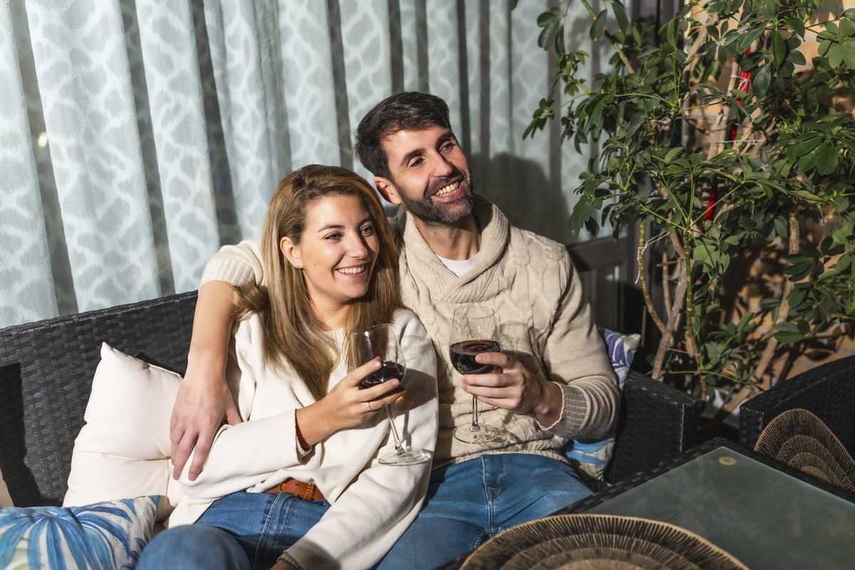 Glückliches Paar, das sich auf einem bequemen Sofa auf der Terrasse entspannt, einen Film ansieht und Rotwein trinkt, um die gemeinsame Freizeit zu genießen Happy couple relaxing on a comfortable sofa ...