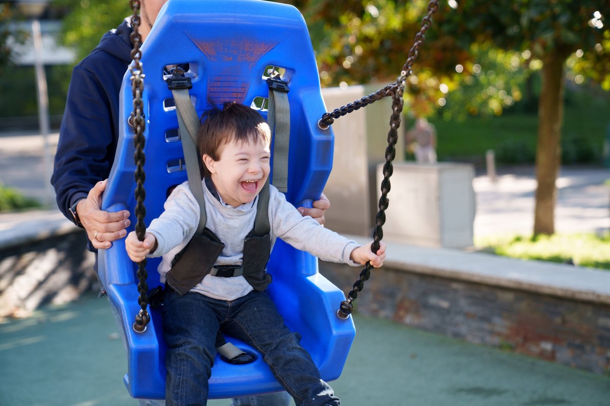A child with Down syndrome happily swings on an adapted swing in a sunny park, supported by an adult. The swing s design ensures safety and inclusion in play Model Released LuisManuelMunoz_ID27747_796 ...