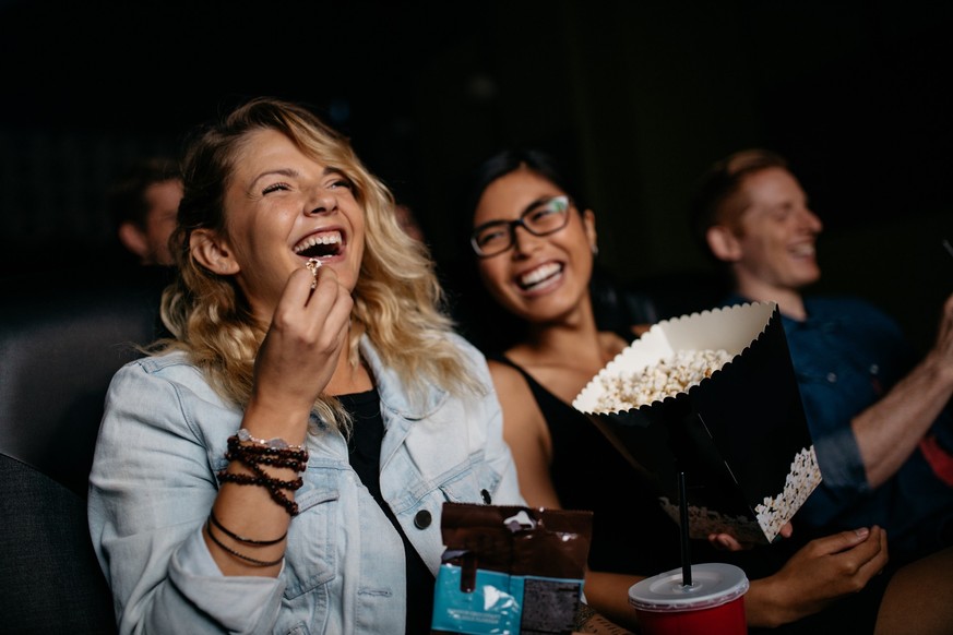 Young woman with friends watching movie in cinema and laughing. Group of people in theater with popcorns and drinks.