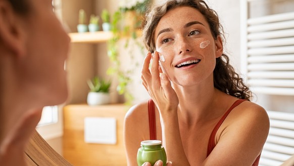 Woman caring of her beautiful skin face standing near mirror in the bathroom. Young woman applying moisturizing cream on her face during morning routine. Smiling natural girl holding little green jar  ...