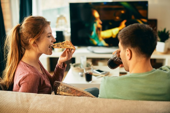 Young couple relaxing while watching TV at home. Focus is on woman eating pizza. Regen Date Zuhause schlechtes Wetter TV Pikcnick Dating