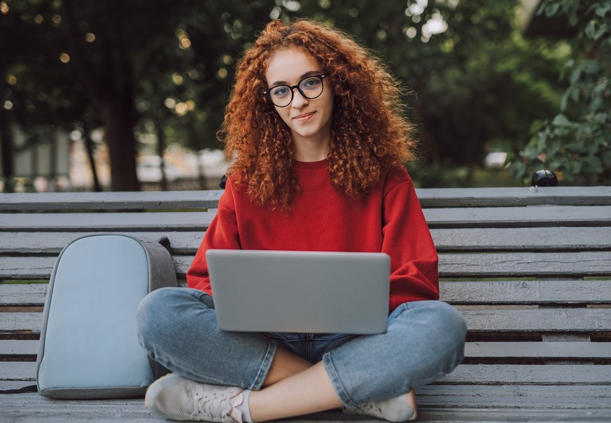 Confident smiling millennial female student with red curly hair wearing casual clothes and eyeglasses using laptop while sitting on bench in city park