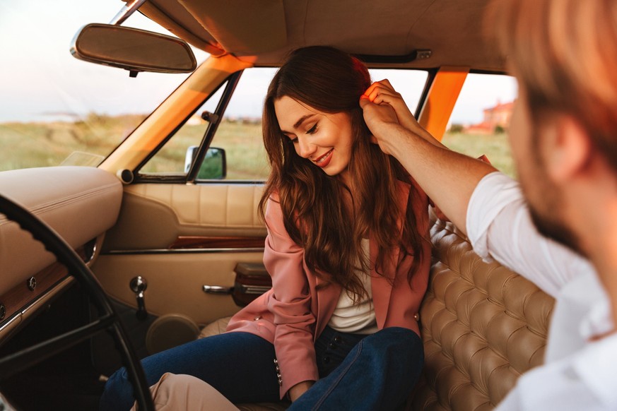 Young man flirting with his smiling girlfriend by touching her hair and holding her hand while sitting inside a retro car