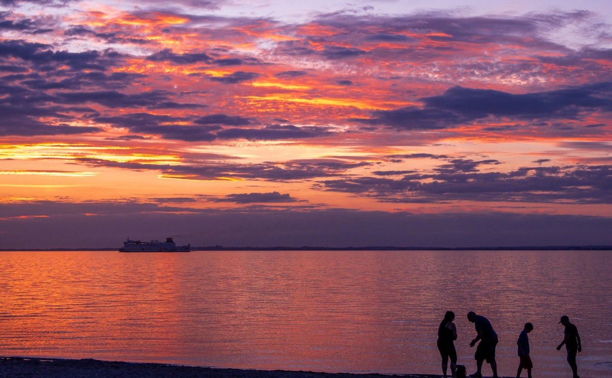 13.08.2023, Mecklenburg-Vorpommern, Gross Schwansee: Eine Familie sammelt Steine während das schwedische Fährschiff "Huckleberry Finn" bei Sonnenuntergang über die Ostsee zum Hafen von Trave ...