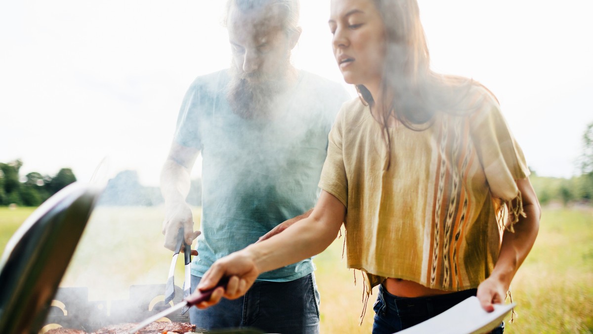A couple cooking food together on a barbecue for their friends at an outdoor lunch on a local farm.